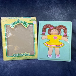 May include: A Cabbage Patch Kids 11-piece plastic puzzle featuring a doll with brown hair, a yellow dress, and pink shoes. The puzzle is packaged in a yellow box with the Cabbage Patch Kids logo.