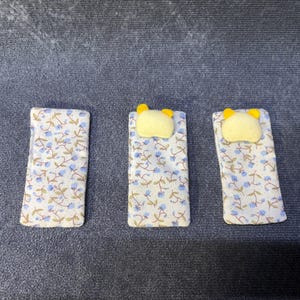 May include: Three miniature doll beds. Each bed has a floral patterned blanket with blue flowers and a yellow pillow. The beds are rectangular and are arranged side by side on a dark gray surface.