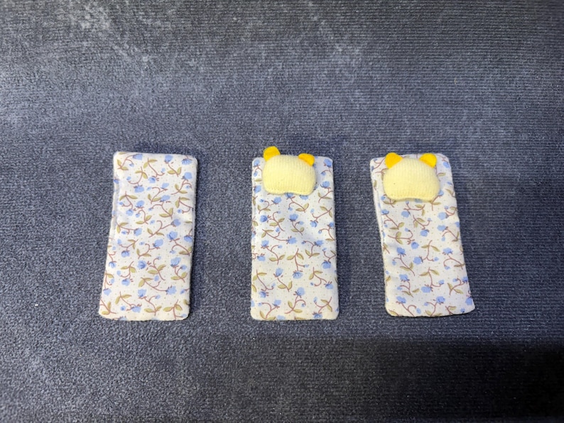 May include: Three miniature doll beds. Each bed has a floral patterned blanket with blue flowers and a yellow pillow. The beds are rectangular and are arranged side by side on a dark gray surface.
