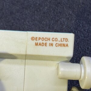 May include: Close-up of a section of a light beige plastic toy. The text "&copy;EPOCH CO.,LTD. MADE IN CHINA" is printed in brown. The toy has a cylindrical knob and a rectangular section.