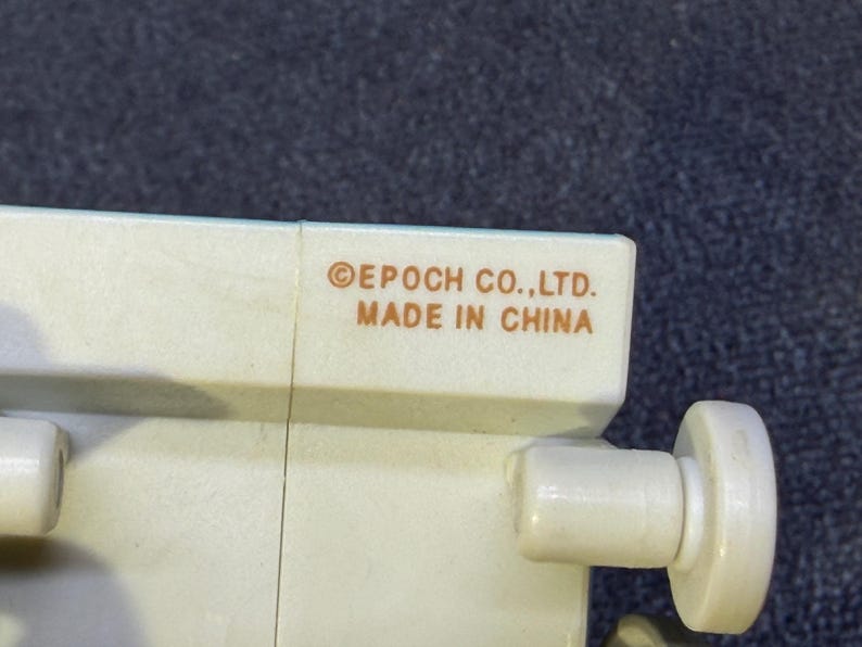 May include: Close-up of a section of a light beige plastic toy. The text "&copy;EPOCH CO.,LTD. MADE IN CHINA" is printed in brown. The toy has a cylindrical knob and a rectangular section.