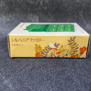 Op de afbeelding: Een kleine, rechthoekige doos met een groene miniatuur dressoir bovenop. De doos is geel met bloemen- en vogelillustraties. Japanse tekst is zichtbaar op de zijkant van de doos.
