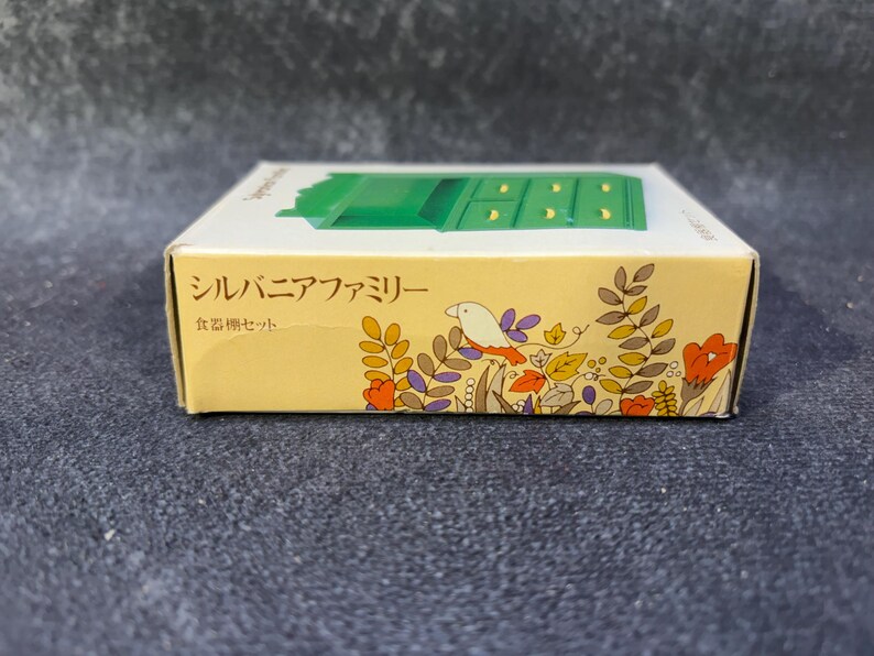 Op de afbeelding: Een kleine, rechthoekige doos met een groene miniatuur dressoir bovenop. De doos is geel met bloemen- en vogelillustraties. Japanse tekst is zichtbaar op de zijkant van de doos.