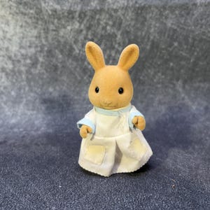 May include: A small, tan rabbit figurine wearing a white coat with blue trim. The rabbit has large, upright ears and small, black eyes. The coat has two small pockets and is buttoned up. The figurine is a toy.