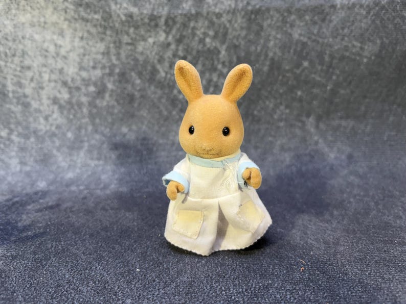 May include: A small, tan rabbit figurine wearing a white coat with blue trim. The rabbit has large, upright ears and small, black eyes. The coat has two small pockets and is buttoned up. The figurine is a toy.