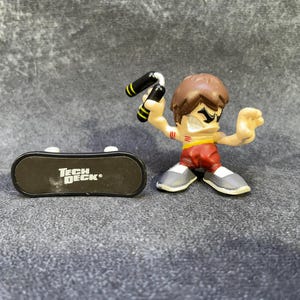 Puede incluir: Un monopatín Tech Deck y una pequeña figura de acción de un artista marcial. La figura tiene cabello castaño, usa pantalones rojos, un cinturón amarillo y zapatos grises. El monopatín es negro con el logo "Tech Deck".