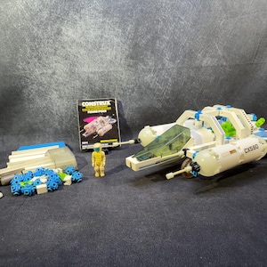 May include: A vintage Construct spacecraft toy set, featuring a white and blue spaceship with a clear cockpit, a yellow astronaut figure, and various building components. The set includes a box with the Construct logo.