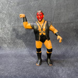 May include: A vintage wrestling action figure with a red and black face, wearing a black vest with silver studs, black trunks, and boots. The figure is posed with one arm raised, showcasing a detailed design.