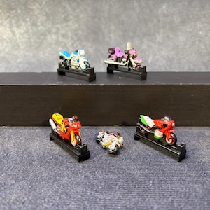 以下が含まれることがあります： 黒いスタンドに飾られた5台のミニチュアバイクのフィギュア。青と白のバイク、ピンクと黒のクルーザー、赤と黄色のスポーツバイク、小さな白と銀のモデル、そして赤、白、緑のスポーツバイクが含まれています。