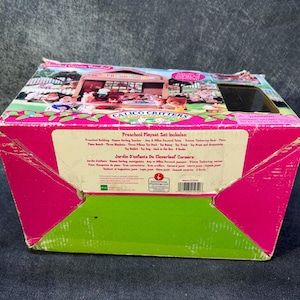 May include: A pink and green Calico Critters Preschool Playset box. The box features an illustration of a schoolhouse with animal figures. Text on the box includes "Preschool Playset Set includes" and "Jardin d'enfants de Cloverleaf Corners".