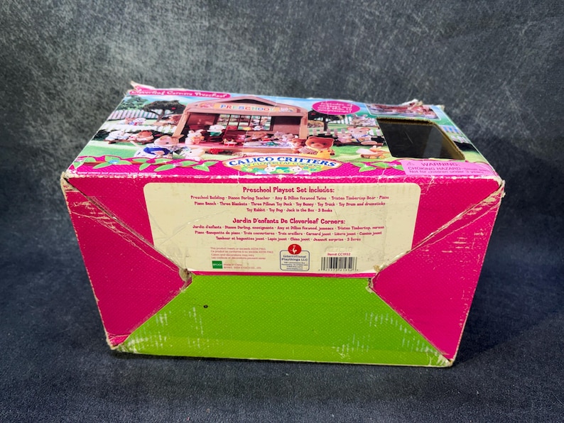May include: A pink and green Calico Critters Preschool Playset box. The box features an illustration of a schoolhouse with animal figures. Text on the box includes "Preschool Playset Set includes" and "Jardin d'enfants de Cloverleaf Corners".