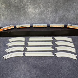 May include: A toy train set consisting of a five-car passenger train with orange, silver, and black coloring, positioned on a long black rectangular base. Below the train are twelve individual plastic track pieces, including straight and curved sections.