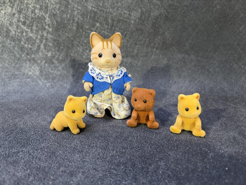 May include: A collection of miniature animal figurines. A cat figure in a blue jacket and floral dress stands with two yellow bear cubs and a brown bear cub. The figurines are made of a soft, flocked material.