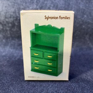 Op de afbeelding: Een groene miniatuur dressoir met goudkleurige handgrepen wordt getoond in de originele kartonnen doos. De doos is gemarkeerd met "Sylvanian Families" en bevat Japanse tekst. De dressoir heeft twee laden boven en drie eronder.