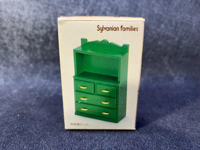 Op de afbeelding: Een groene miniatuur dressoir met goudkleurige handgrepen wordt getoond in de originele kartonnen doos. De doos is gemarkeerd met "Sylvanian Families" en bevat Japanse tekst. De dressoir heeft twee laden boven en drie eronder.