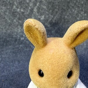 May include: A close-up of a tan rabbit figurine with large, upright ears and black eyes. The rabbit has a soft, fuzzy texture and is wearing a white garment. The background is a textured, dark gray surface.