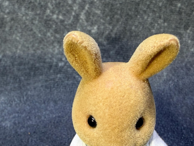 May include: A close-up of a tan rabbit figurine with large, upright ears and black eyes. The rabbit has a soft, fuzzy texture and is wearing a white garment. The background is a textured, dark gray surface.