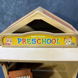 May include: A tan-colored toy preschool building with a brown roof. The sign reads "Main Street PRESCHOOL" in blue and red letters, with cartoon animal illustrations on each side. A toy piano is visible inside.