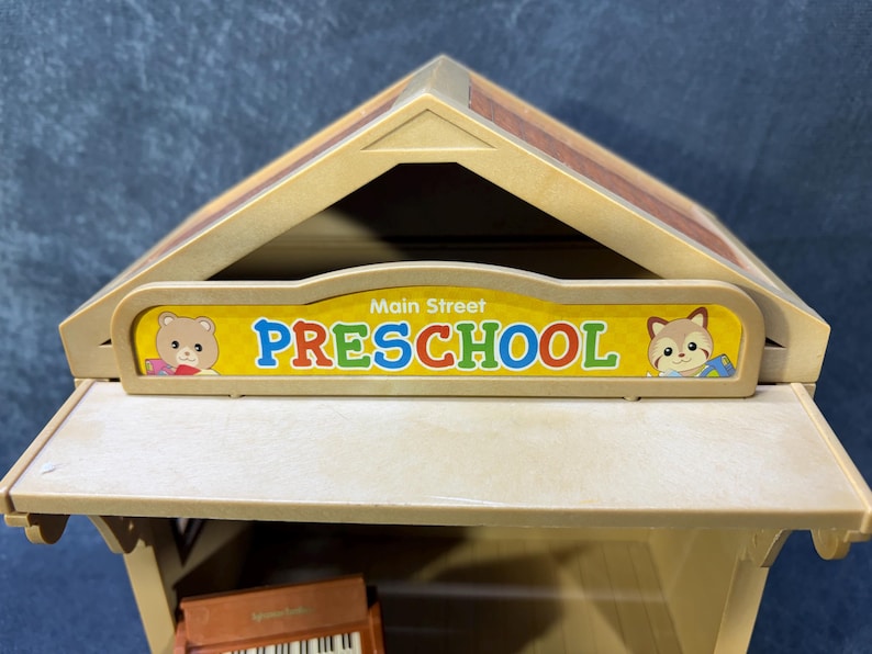 May include: A tan-colored toy preschool building with a brown roof. The sign reads "Main Street PRESCHOOL" in blue and red letters, with cartoon animal illustrations on each side. A toy piano is visible inside.