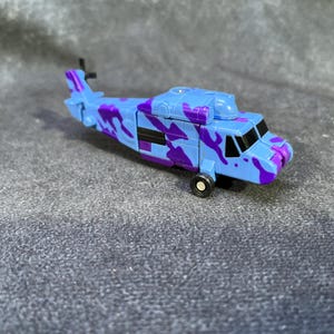 Puede incluir: Un helicóptero de juguete azul y morado con camuflaje, con ventanas negras y un rotor negro. El juguete tiene una sola rueda negra y está sobre un fondo gris. El helicóptero es un juguete.