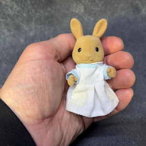 May include: A small, tan rabbit figurine wearing a white dress with a light blue collar and sleeves. The rabbit has black eyes and upright ears. The figurine is held in a person's hand against a dark gray background.