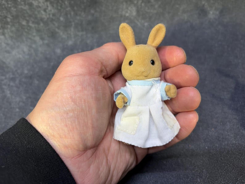 May include: A small, tan rabbit figurine wearing a white dress with a light blue collar and sleeves. The rabbit has black eyes and upright ears. The figurine is held in a person's hand against a dark gray background.