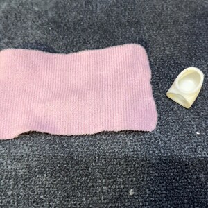 May include: A light pink, rectangular fabric item and a small, off-white plastic object. The fabric appears to be a knitted material. The plastic object has a curved shape with a circular indentation.