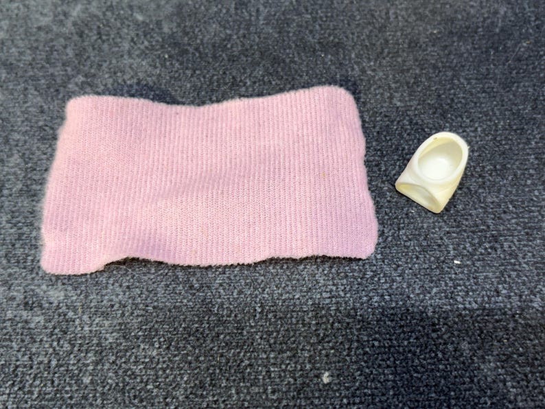 May include: A light pink, rectangular fabric item and a small, off-white plastic object. The fabric appears to be a knitted material. The plastic object has a curved shape with a circular indentation.