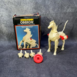 May include: A cream-coloured Micronauts Oberon horse action figure with red accents, standing next to its original cardboard packaging box. The box features the text Micronauts Oberon. Four small cream-coloured components and one red circular piece are included.