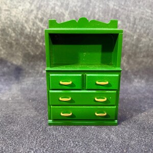 Op de afbeelding: Een miniatuur groene houten dressoir met vier lades en goudkleurige handgrepen. Het bovenste gedeelte heeft een plank. Het dressoir is geschilderd in een levendige groene kleur en het algehele ontwerp is eenvoudig en klassiek.