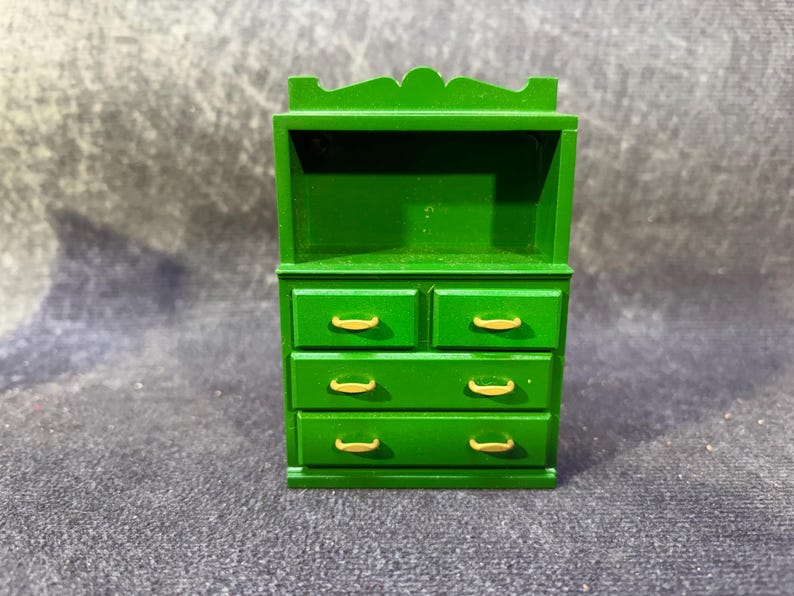 Op de afbeelding: Een miniatuur groene houten dressoir met vier lades en goudkleurige handgrepen. Het bovenste gedeelte heeft een plank. Het dressoir is geschilderd in een levendige groene kleur en het algehele ontwerp is eenvoudig en klassiek.