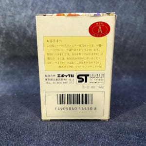 Op de afbeelding: Een kleine, rechthoekige doos met Japanse tekst en een rode ronde sticker met de letter "A". De doos is wit met een barcode en productinformatie erop gedrukt. De doos staat op een blauw oppervlak.