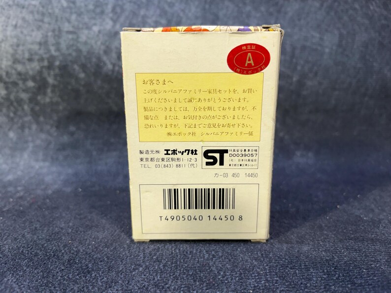 Op de afbeelding: Een kleine, rechthoekige doos met Japanse tekst en een rode ronde sticker met de letter "A". De doos is wit met een barcode en productinformatie erop gedrukt. De doos staat op een blauw oppervlak.