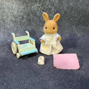May include: A miniature toy set featuring a brown rabbit figure in a white and blue outfit, a light blue and white wheelchair, a small white shoe, and a pink blanket. The toys are designed for children.
