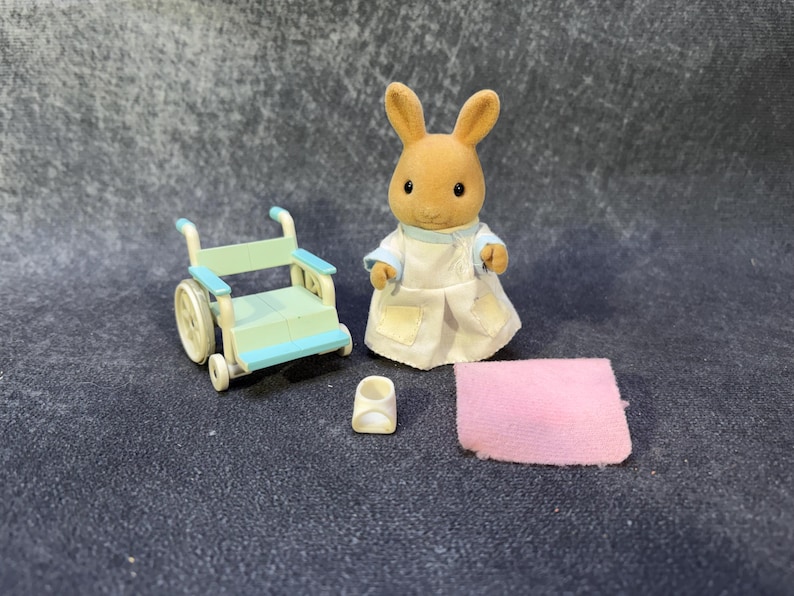 May include: A miniature toy set featuring a brown rabbit figure in a white and blue outfit, a light blue and white wheelchair, a small white shoe, and a pink blanket. The toys are designed for children.