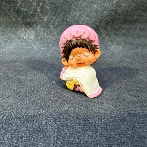 May include: A small figurine of a sleeping child wearing a pink bonnet and a white gown with pink trim. The child has brown hair and a peach-colored face. The figurine is resting on a dark gray surface.