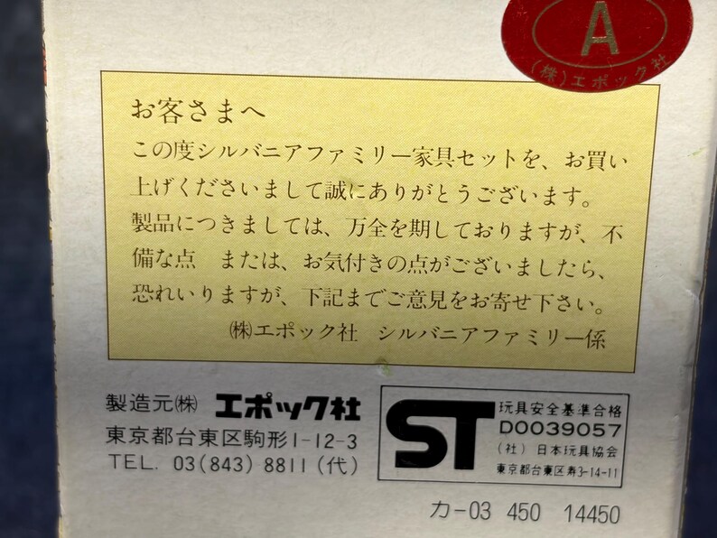 Op de afbeelding: Een close-up van een doos met Japanse tekst en een rode ronde sticker. De tekst staat op een gele achtergrond en bevat de informatie van de fabrikant en een veiligheidsmerk.