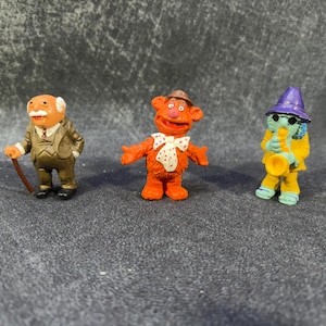 Könnte beinhalten: Drei kleine Figuren von Muppet-Charakteren. Einer ist ein älterer Mann mit einem Stock, ein anderer ist ein orangefarbener Bär mit einer Fliege, und der dritte ist eine blaue Figur, die Saxophon spielt und einen lila Hut trägt.