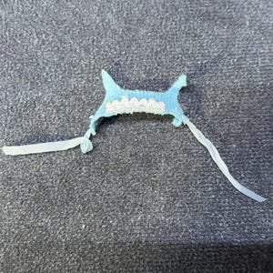 May include: A small, light blue fabric item with a scalloped white lace trim. Two white ribbons extend from the sides. The item is likely a decorative piece or part of a garment, possibly for a baby or doll.