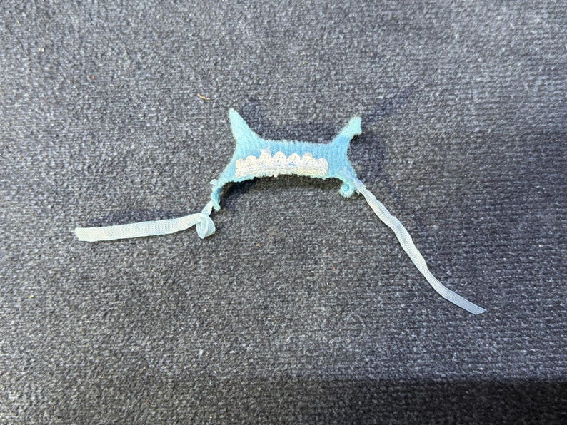 May include: A small, light blue fabric item with a scalloped white lace trim. Two white ribbons extend from the sides. The item is likely a decorative piece or part of a garment, possibly for a baby or doll.