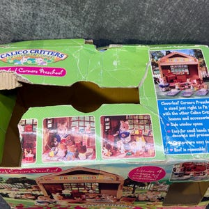 May include: A green cardboard box for a Calico Critters Cloverleaf Corners Preschool playset. The box features images of the playset and text that reads "Cloverleaf Corners Preschool" and "Includes 4 Critters and more accessories!"