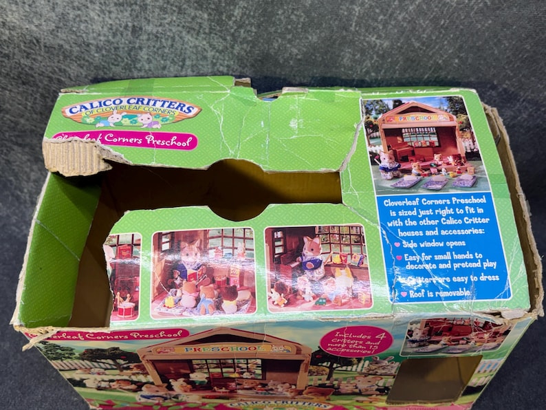 May include: A green cardboard box for a Calico Critters Cloverleaf Corners Preschool playset. The box features images of the playset and text that reads "Cloverleaf Corners Preschool" and "Includes 4 Critters and more accessories!"