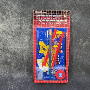 May include: A vintage Transformers school kit in its original packaging. The red and blue package contains a ruler, pencil, eraser, and other school supplies. The Transformers logo is prominently displayed.