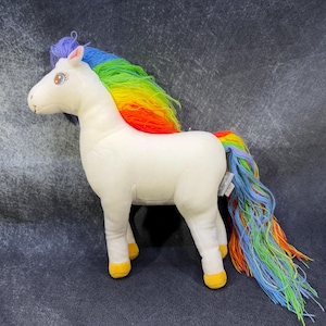 May include: A white plush unicorn toy with a rainbow mane and tail. The mane transitions from purple to blue, green, yellow, and red. The tail mirrors the colours of the mane. The unicorn has yellow hooves and a stitched face.
