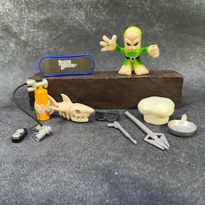 Puede incluir: Colección de juguetes en miniatura, que incluye una figura alienígena verde y amarilla, una patineta Tech Deck, un esqueleto de tiburón, equipo de buceo y utensilios de cocina. Los juguetes están dispuestos sobre un bloque de madera oscura.