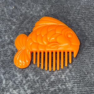 May include: An orange plastic fish-shaped comb. The comb features a detailed fish design with fins and scales. The comb's teeth are vertical and evenly spaced.