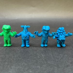 Puede incluir: Cuatro pequeñas figuras de juguete de plástico azul y verde con diferentes diseños. Las figuras están de pie sobre una superficie gris.