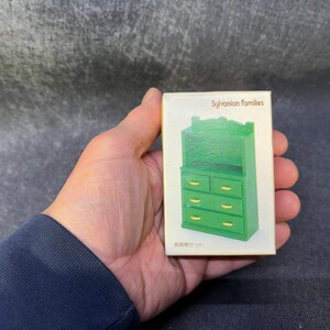 Op de afbeelding: Een groene miniatuurkast met lades en een bovenste plank, onderdeel van de Sylvanian Families-collectie. De kast wordt gepresenteerd op een witte doos, met tekst in het Engels en Japans. De kast heeft goudkleurige ladeknopen.