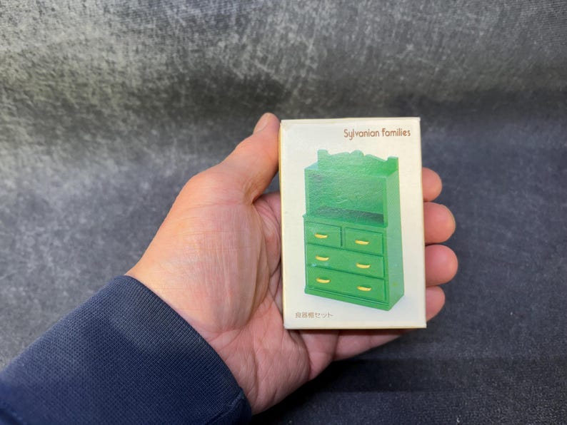 Op de afbeelding: Een groene miniatuurkast met lades en een bovenste plank, onderdeel van de Sylvanian Families-collectie. De kast wordt gepresenteerd op een witte doos, met tekst in het Engels en Japans. De kast heeft goudkleurige ladeknopen.
