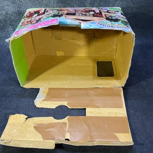 May include: An open, partially assembled cardboard box with a colorful printed top. The box is tan with green and brown accents. A small plastic bag of colorful beads sits to the right of the box.